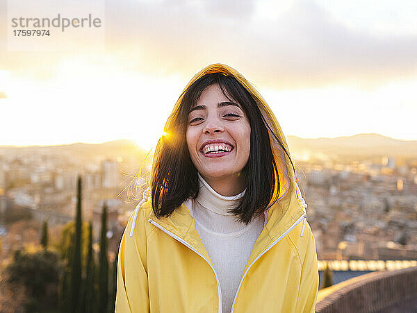Lächelnde junge Frau im gelben Regenmantel mit Stadt im Hintergrund bei Sonnenuntergang