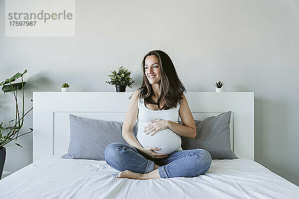Lächelnde schwangere Frau sitzt mit gekreuzten Beinen zu Hause im Bett