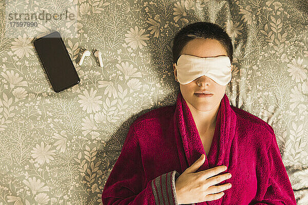 Woman wearing eye mask lying on bed by mobile phone and wireless headphones at home