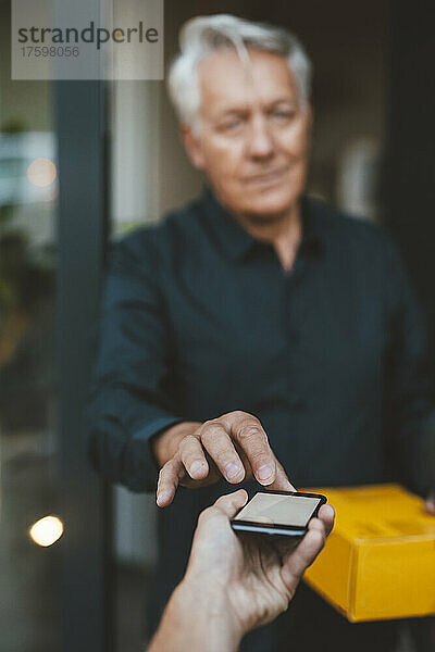 Älterer Mann mit Paketsignatur auf Smartphone  das vom Zusteller gehalten wird