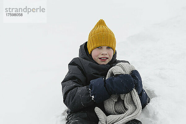 Junge mit gelber Strickmütze und grauem Schal sitzt auf Schnee