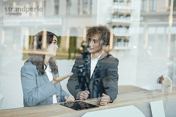 Junge Geschäftsfrau diskutiert mit Geschäftsmann am Schreibtisch im Coworking-Büro  gesehen durch Glas