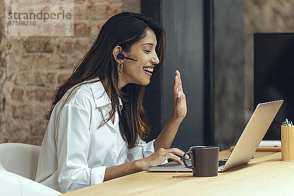 Glückliche Geschäftsfrau winkt bei Videoanruf im Coworking-Büro mit der Hand