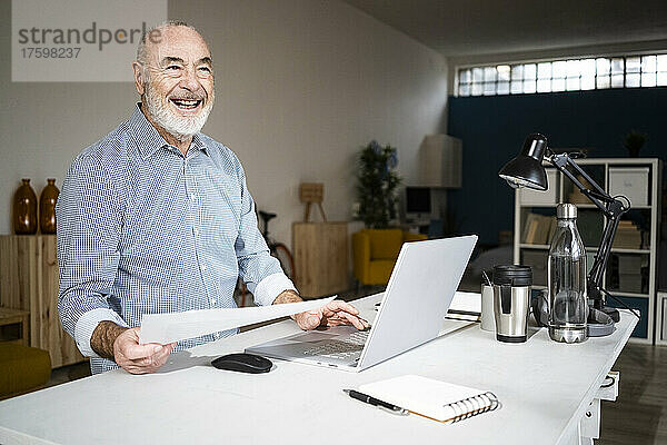 Fröhlicher Geschäftsmann mit Dokument und Laptop am Schreibtisch