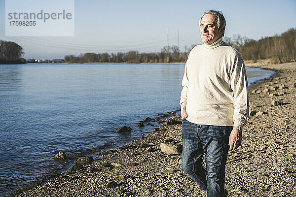 Lächelnder älterer Mann mit der Hand in der Tasche  der am Strand spaziert