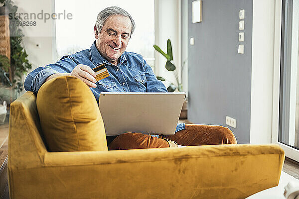 Lächelnder älterer Mann mit Laptop und Kreditkarte sitzt auf dem Sofa im Wohnzimmer