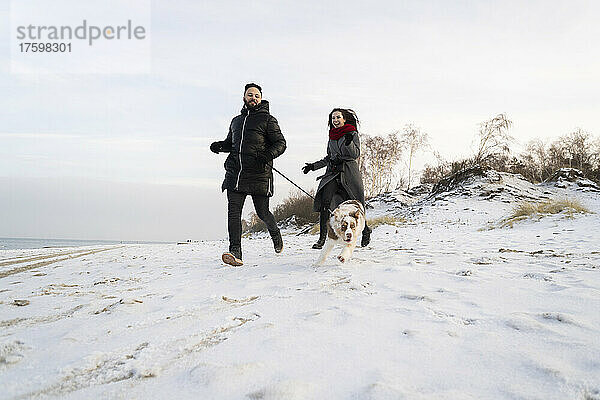 Junges Paar läuft bei Sonnenuntergang mit Hund im Schnee