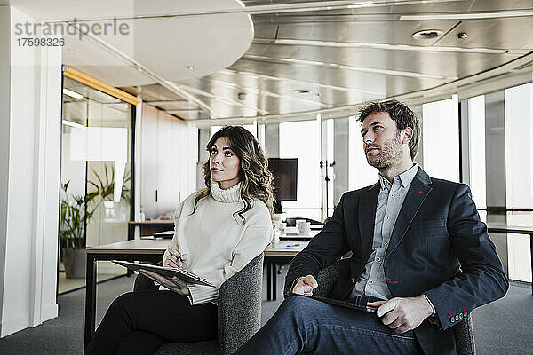 Geschäftsfrau mit Klemmbrett sitzt neben Geschäftsmann mit digitalem Tablet im Sitzungssaal