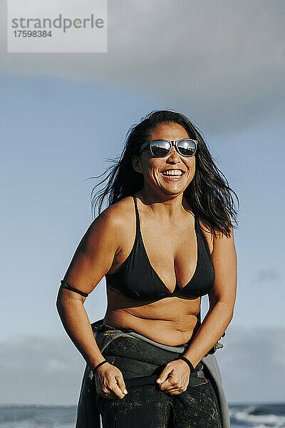 Glückliche Frau in Neoprenanzug und Bikini am Strand