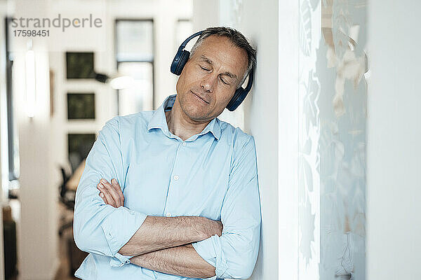 Geschäftsmann mit geschlossenen Augen  der Musik hört und sich im Büro an die Wand lehnt