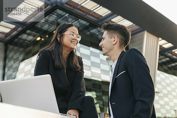 Lächelnde Geschäftsfrau mit Laptop blickt Kollegin vor Bürogebäude an