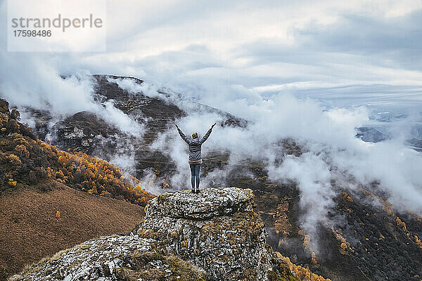 Female hiker standing with raised arms on top of outcrop in autumn mountains of North Caucasus