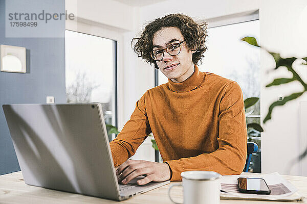 Junger Mann mit Laptop sitzt am Tisch im Wohnzimmer