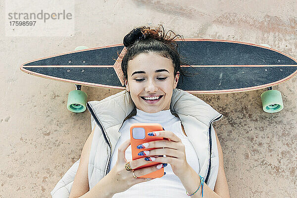 Lächelnde Frau liegt auf Skateboard und benutzt Smartphone im Park