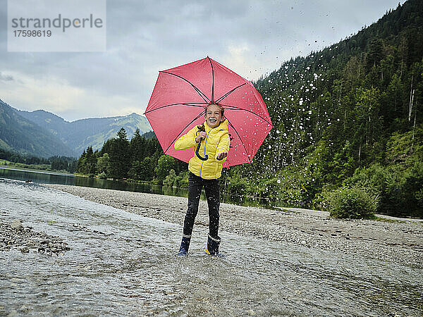 Fröhliches Mädchen mit Regenschirm genießt das Wasserspritzen am Seeufer