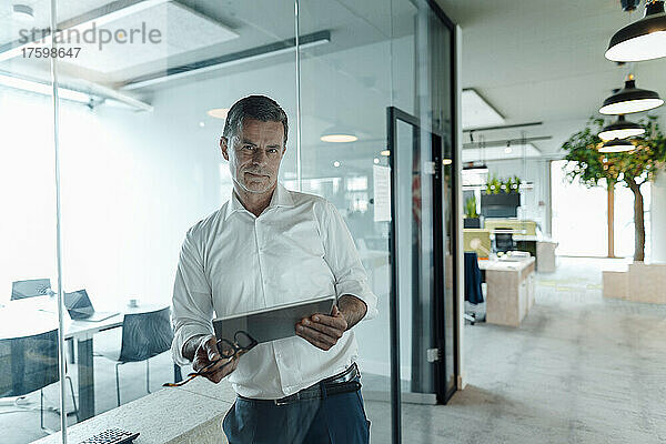 Selbstbewusster Geschäftsmann mit Tablet-PC  der im Büro an einer Glaswand lehnt