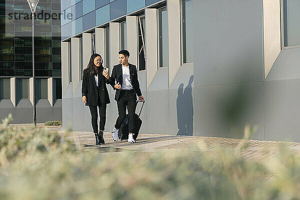 Businessman with trolley bag walking with colleague on footpath by office building
