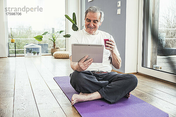 Lächelnder älterer Mann sitzt mit gekreuzten Beinen auf einer Trainingsmatte und schaut sich zu Hause Videos über einen Tablet-PC an