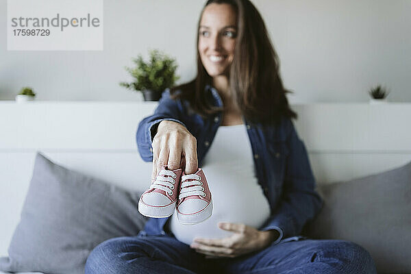 Smiling pregnant woman showing baby booties in bedroom at home
