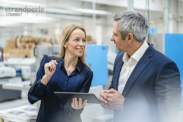 Glückliche blonde Geschäftsfrau hält Tablet-PC in der Hand und diskutiert mit Kollegen in der Fabrik