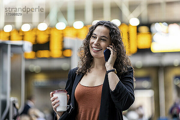 Lächelnde Frau mit Einwegkaffeetasse  die auf dem Smartphone spricht