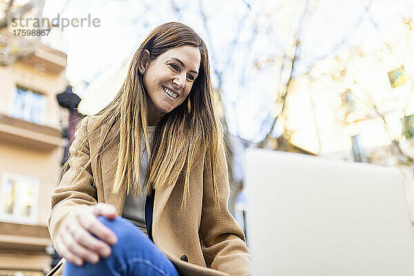 Lächelnde Frau mit braunen Haaren  die in der Stadt auf ihren Laptop schaut