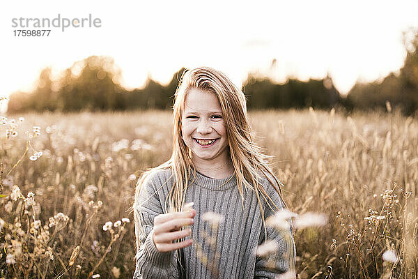 Fröhliches blondes Mädchen hält bei Sonnenuntergang eine Blume auf dem Feld