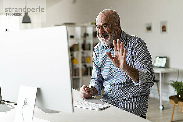 Cheerful senior businessman waving hand on video call through desktop computer in home office