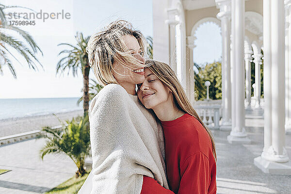 Lächelndes Teenager-Mädchen mit geschlossenen Augen umarmt Mutter an einem sonnigen Tag