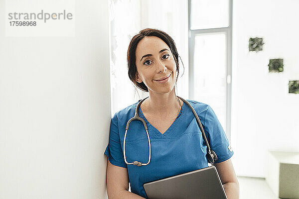 Lächelnder Arzt mit Tablet-PC lehnt an der Wand im Krankenhaus