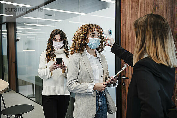 Geschäftsfrau überprüft Temperatur eines Kollegen  der im Büro eine schützende Gesichtsmaske trägt