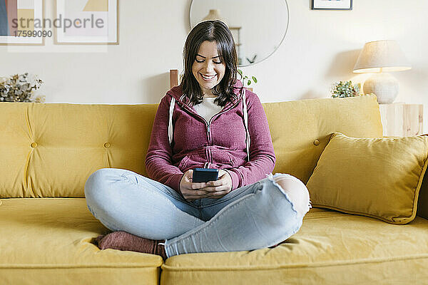 Lächelnde junge Frau  die zu Hause auf dem Sofa mit dem Handy im Internet surft