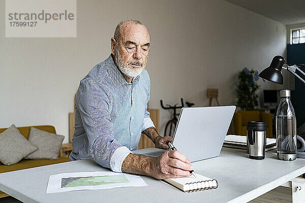 Geschäftsmann schreibt in Notizblock und sitzt mit Laptop am Schreibtisch