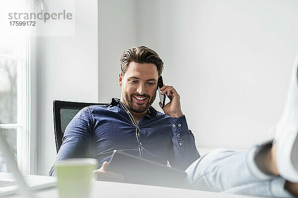 Lächelnder Geschäftsmann mit Tablet-PC  der im Büro auf dem Smartphone spricht