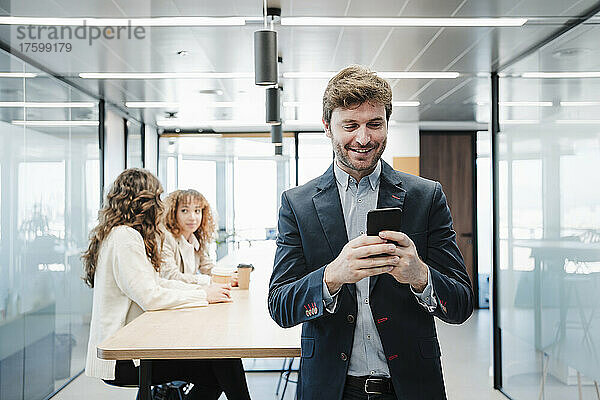 Lächelnder Geschäftsmann  der im Büro Textnachrichten über sein Smartphone sendet