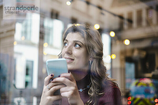 Nachdenkliche Frau mit Smartphone  gesehen durch das Fenster eines Cafés
