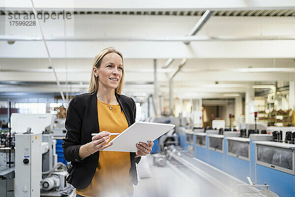 Lächelnde blonde Geschäftsfrau mit Tablet-PC steht in der Fabrik