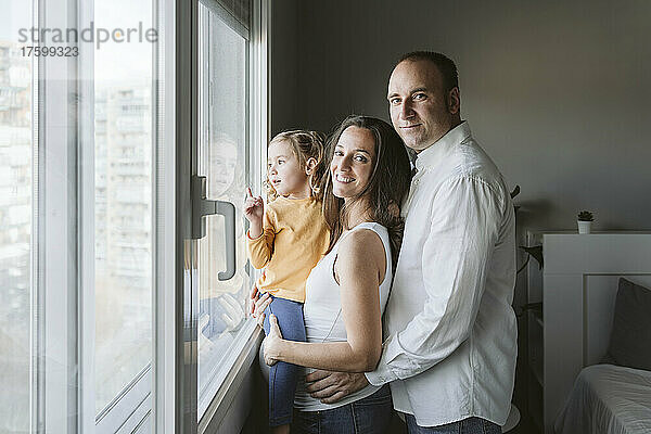 Lächelnde Eltern stehen mit ihrer Tochter und schauen zu Hause durch das Fenster