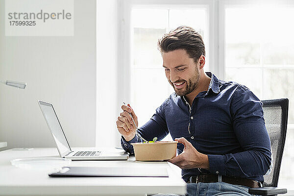 Lächelnder Geschäftsmann  der Salat am Schreibtisch im Büro isst