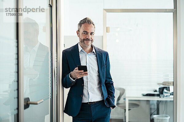 Lächelnder Geschäftsmann lehnt mit der Hand in der Tasche und hält sein Smartphone im Büro