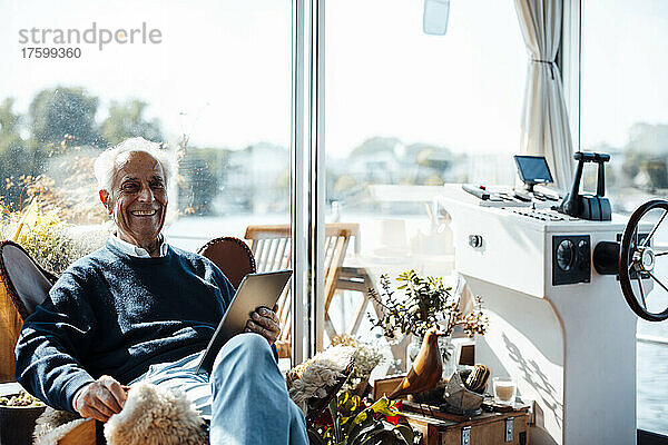 Fröhlicher älterer Mann mit Tablet-PC sitzt am Hausboot