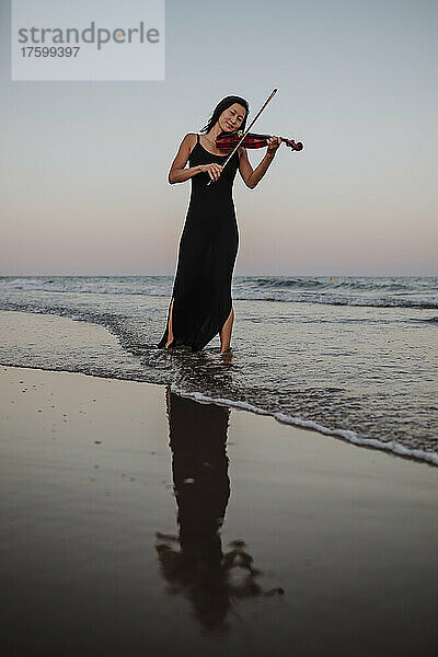 Frau spielt Geige und steht bei Sonnenuntergang im Meer