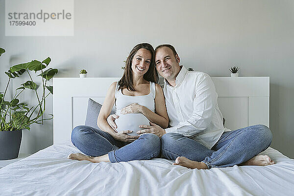 Glücklicher Mann sitzt mit schwangerer Frau zu Hause im Bett