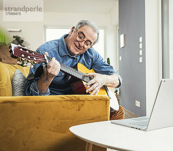 Älterer Mann mit Brille spielt Gitarre auf Sofa im Wohnzimmer