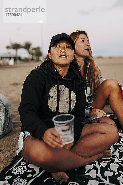Nachdenkliche Frau mit Bierbecher sitzt neben einer Freundin am Strand