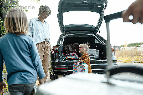 Mutter blickt im Urlaub Tochter mit Koffer am Kofferraum eines Autos an