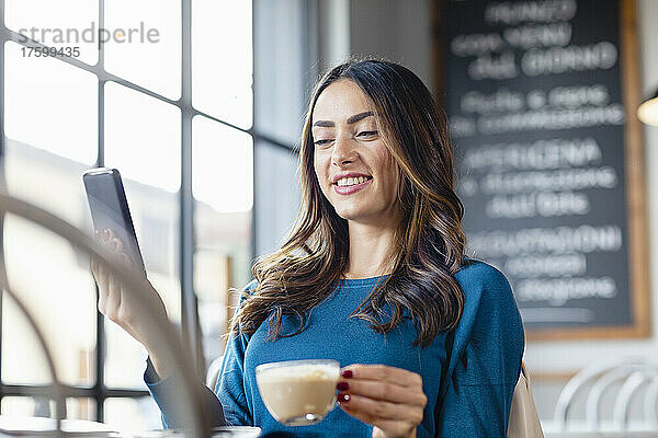 Lächelnde Frau mit Kaffeetasse bei Videoanruf über Mobiltelefon im Café