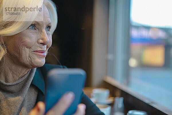 Ältere Frau mit Smartphone schaut aus dem Fenster