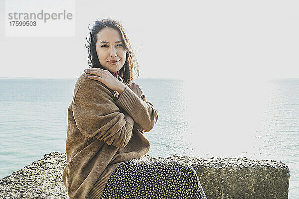 Schöne junge Frau umarmt sich selbst und sitzt auf einem Felsen am Strand