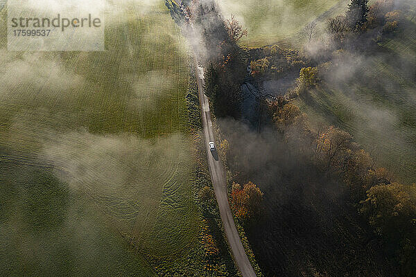 Drone view of countryside dirt road stretching along river at foggy autumn morning